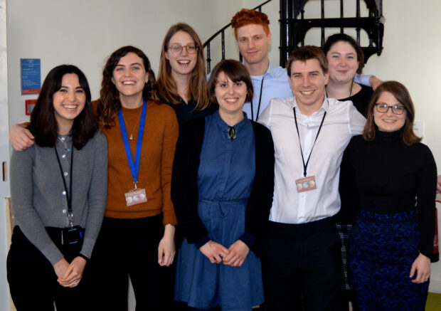 A smiley group photo of eight of the Open Innovation Team's PhD students. From left to right: Sarah Oufan, Rosa Hodgkin, Sophia Peacock, Lizzie Atkinson, Marc Goldfinger, Peter West, Sarah Sheehy, and Rachel Efrat.