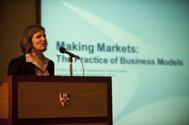 The image shows Katy Mason, the author of this blog post, giving a talk with her presentation behind her which reads 'Making Markets: The Practise of Business Models'